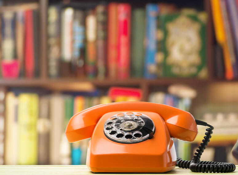 A photo of an orange rotary dial telephone in front of library book shelves