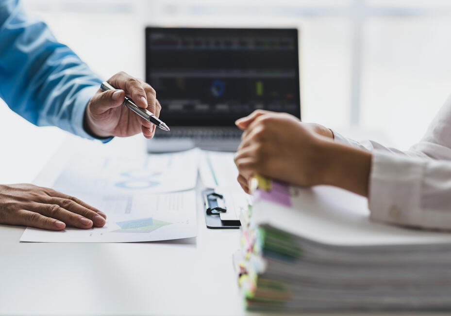 Image of one person's hands on top of a stack of papers across from another person's hands holding a pen pointing at charts